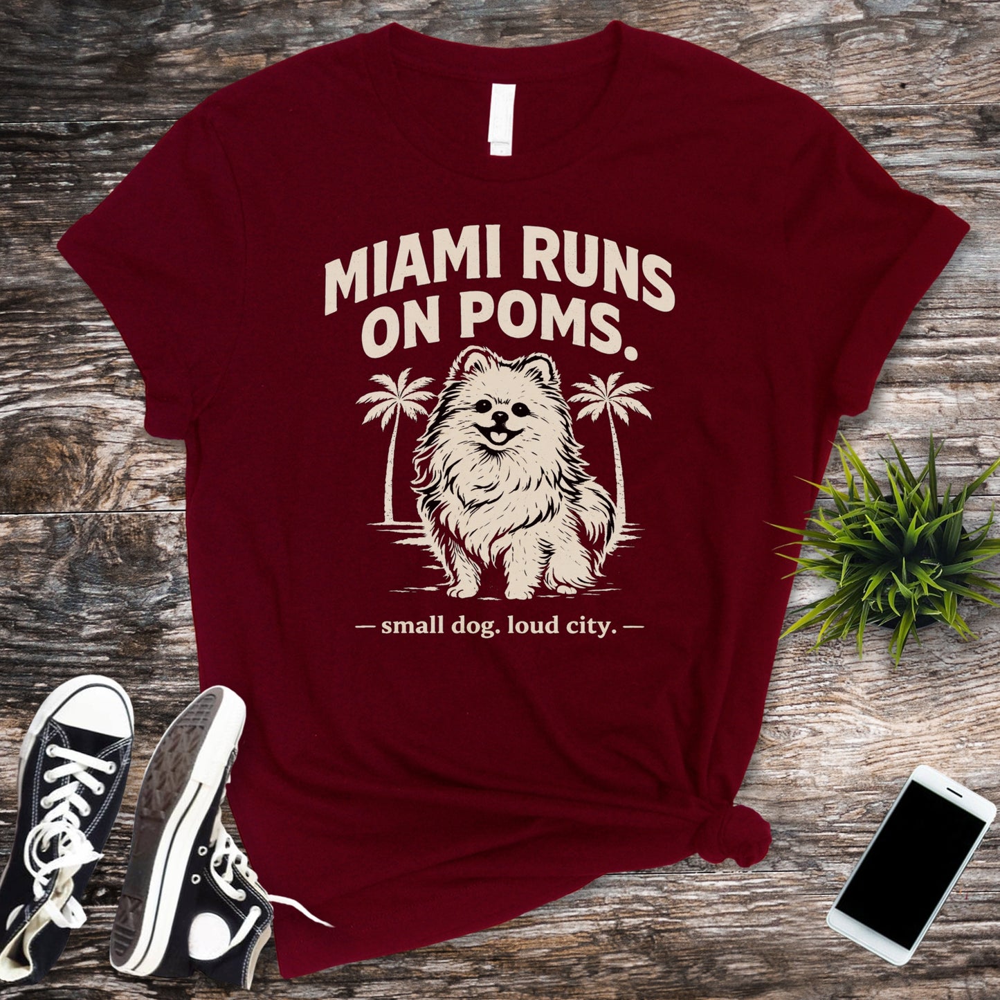 Maroon t-shirt with dog graphic and text on a wooden surface with sneakers and a phone.