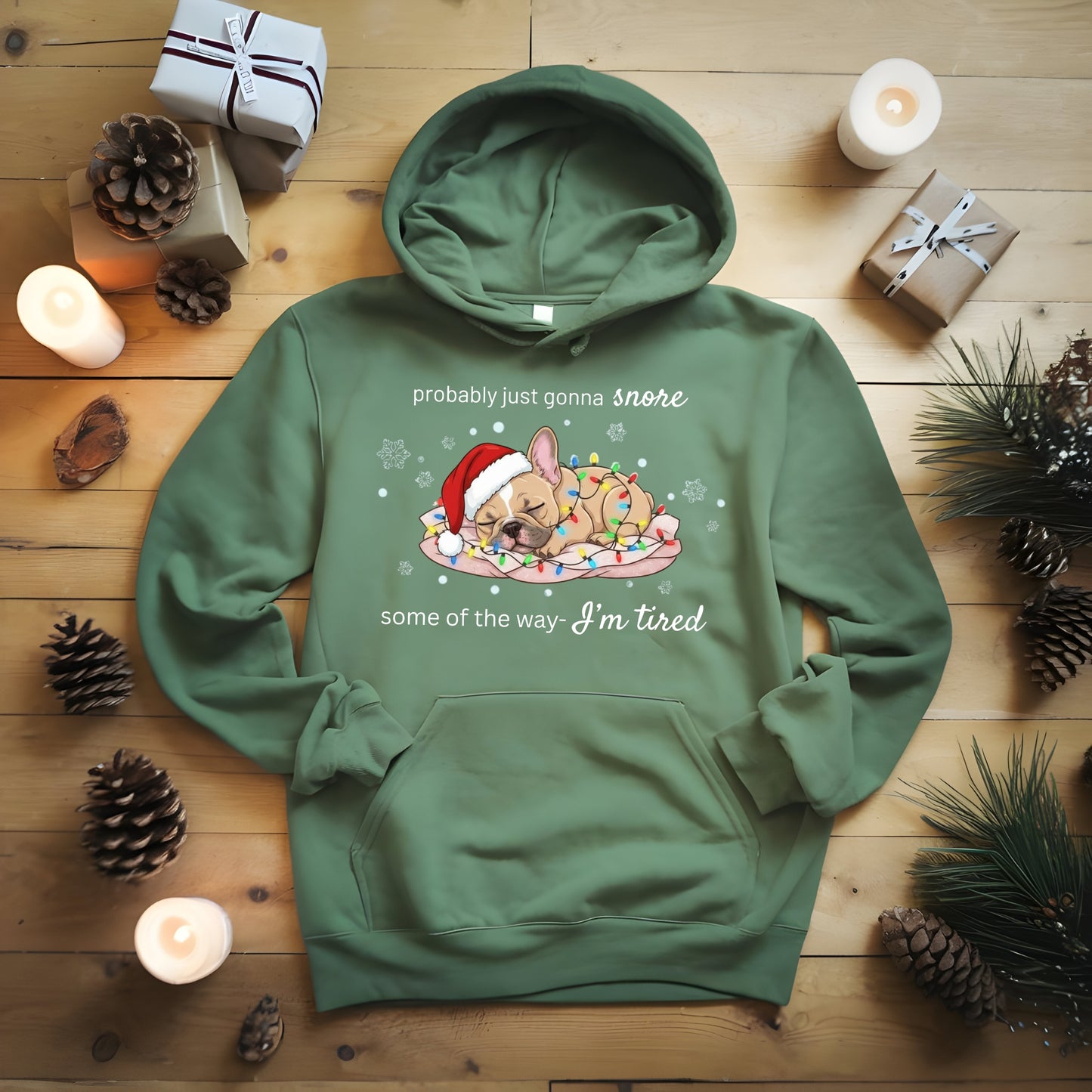 Green hoodie with a Christmas-themed design on a wooden surface with candles and pinecones.