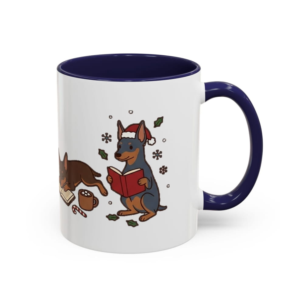 White ceramic mug with a navy interior and navy handle, showing a black-and-tan Mini Pinscher in a Santa hat reading a red book, with another Min Pin lying beside it.