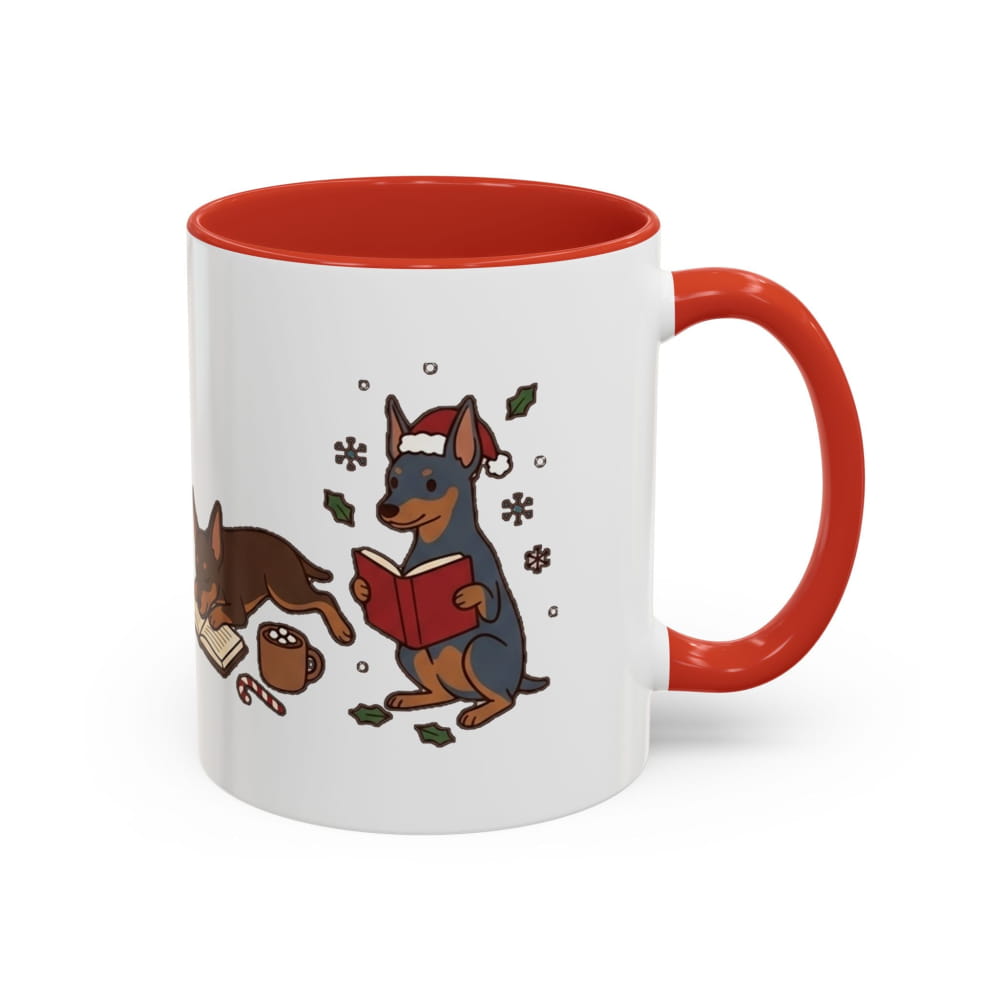 White ceramic mug with a red interior and red handle, showing a black-and-tan Mini Pinscher in a Santa hat reading a red book, with another Min Pin lying beside it.