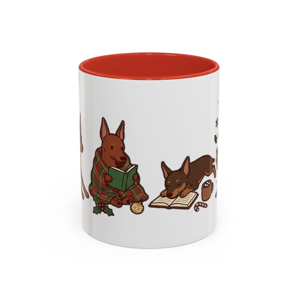 White ceramic mug with a red interior, showing a red Min Pin in a plaid blanket reading a green book beside a dark Min Pin lying on an open book.