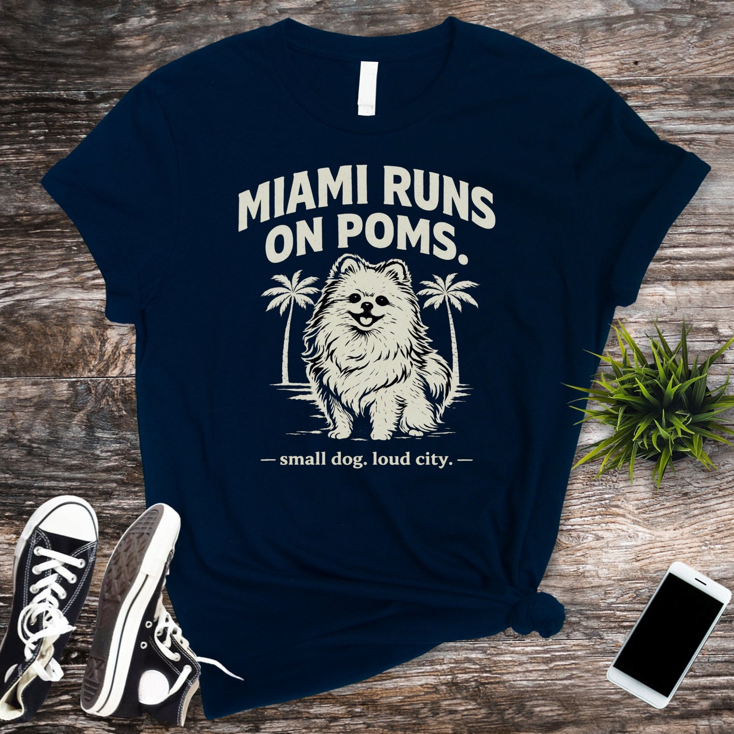 Navy blue t-shirt with dog graphic and text on a wooden surface with sneakers and a phone.