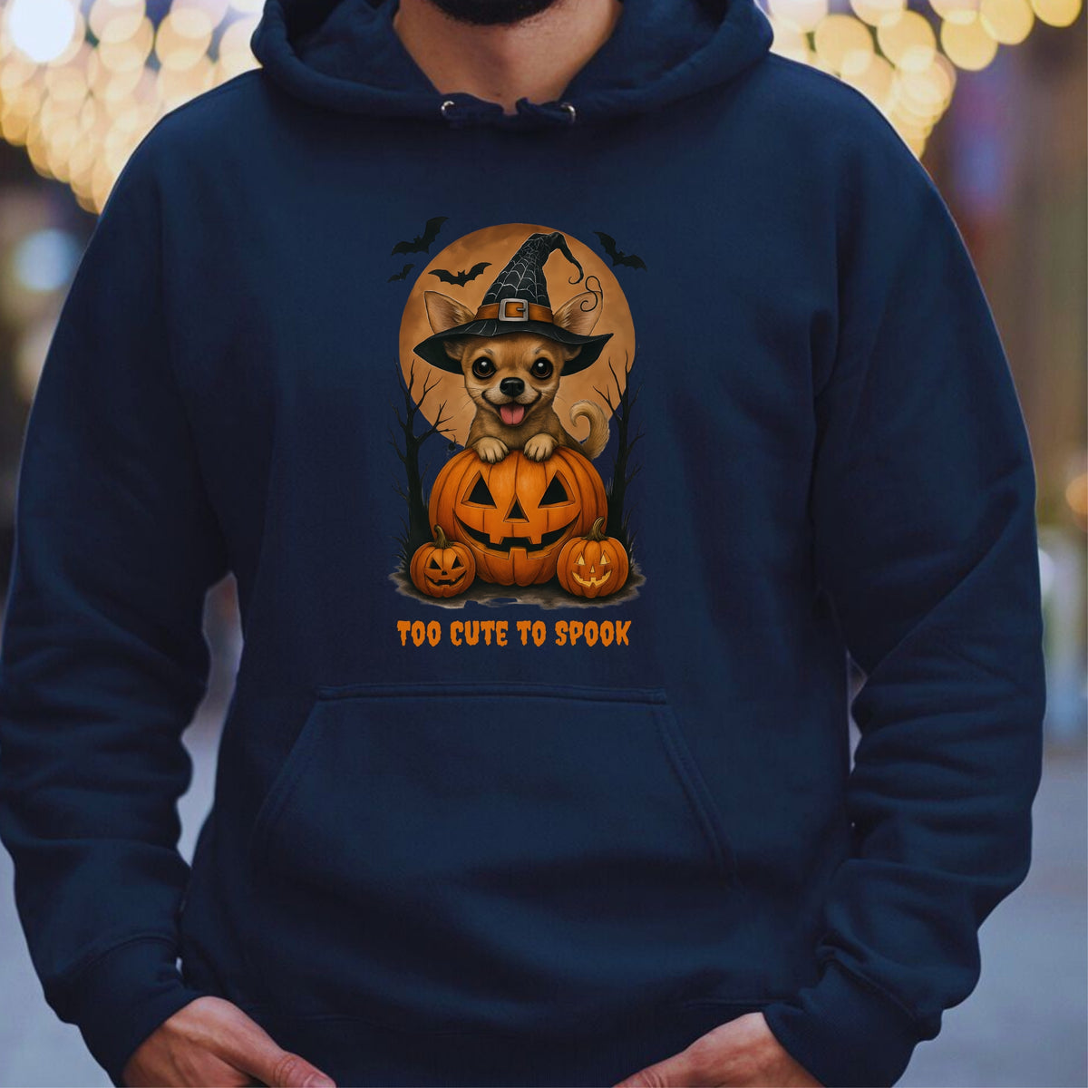 A person wearing a navy blue hoodie with a Halloween graphic of a chihuahua in a witch hat sitting on jack-o'-lantern pumpkins, with the text 'TOO CUTE TO SPOOK' below, against a blurred background with warm lights.