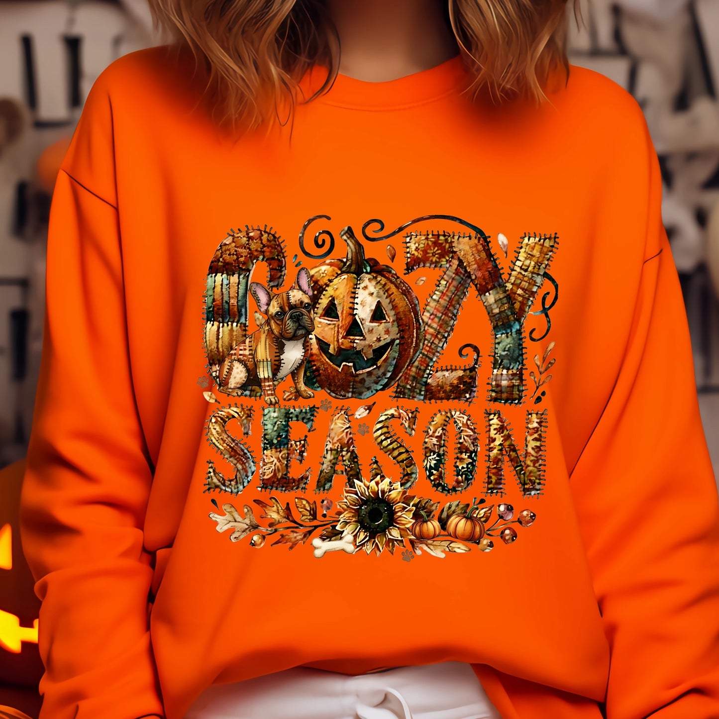 Person wearing an orange sweatshirt with ' cozy season' design featuring pumpkins and leaves.