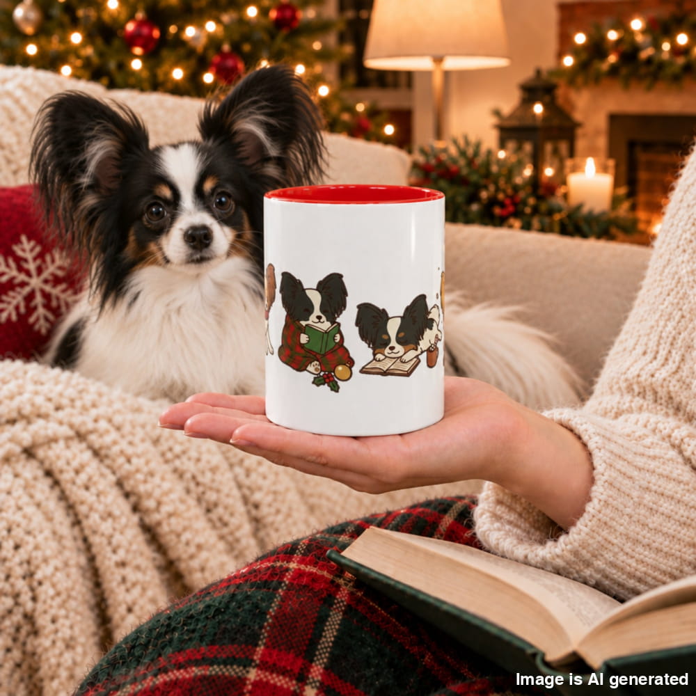 A hand holds a white ceramic mug with a red interior and a Christmas illustration of Papillons reading, while a real Papillon rests on a cozy couch.