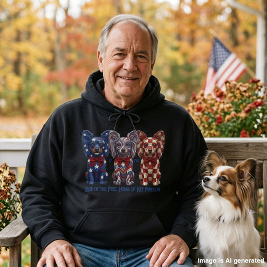 An older man wearing a black hoodie with a patriotic bear graphic sits on a wooden bench outdoors next to a Papillon dog. The background shows a blurred autumn garden with an American flag.