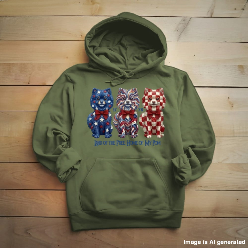An olive green pullover hoodie laid flat on a light wood plank background. The front of the hoodie features a graphic of three small dogs decorated in patriotic red, white, and blue patterns, with the text 'Kind of the Free Dogs of NY' printed below.