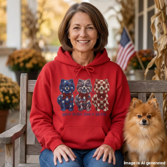 A smiling woman sits on a wooden porch bench wearing a bright red hoodie with a patriotic dog graphic and blue jeans, with a small fluffy dog beside her. The background shows a softly blurred porch setting with autumn decor and an American flag.