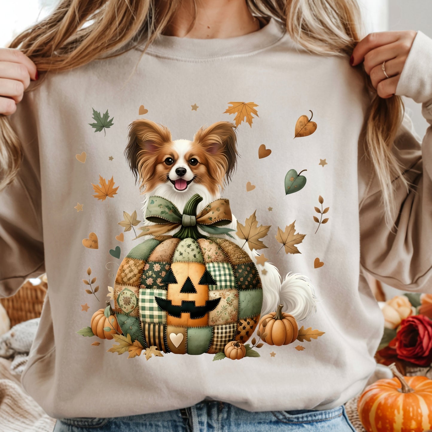Person wearing a beige sweatshirt with a dog in a pumpkin patch design, surrounded by autumn leaves and pumpkins.