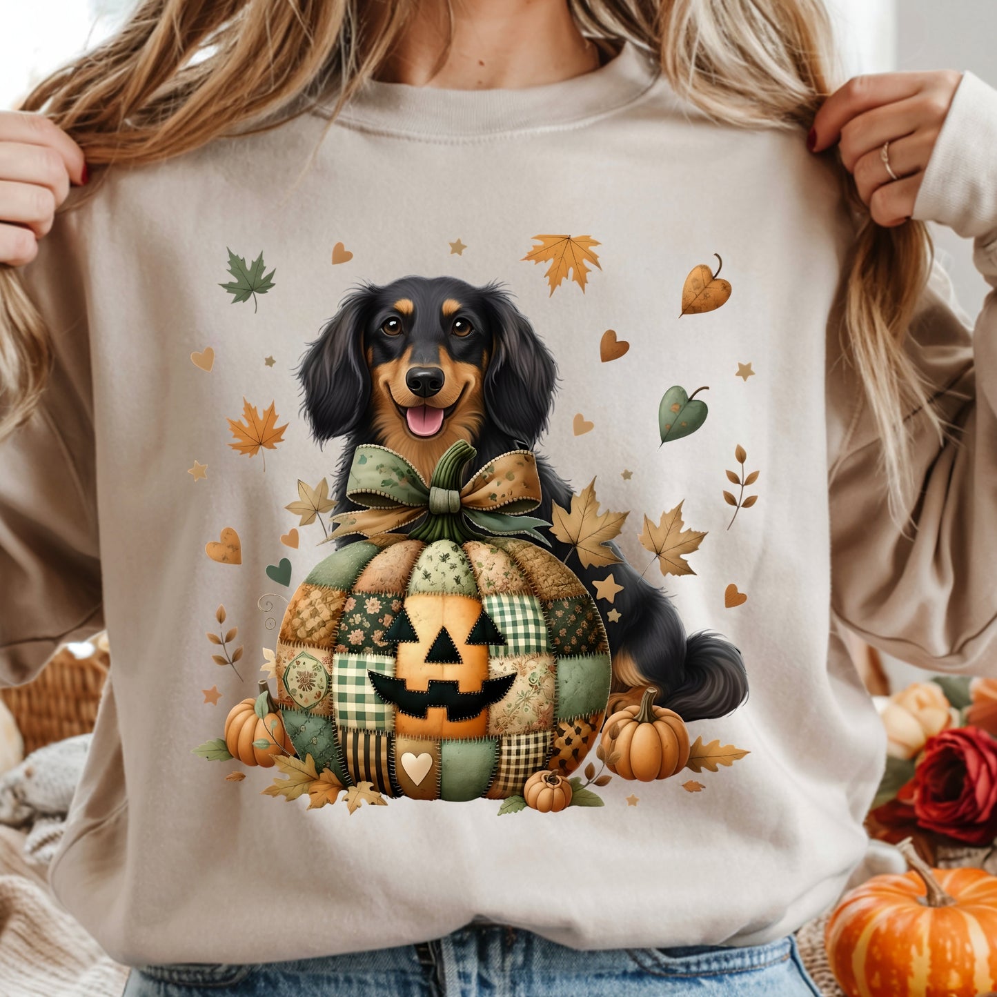 Person wearing a beige sweatshirt with a dachshund and pumpkin design, surrounded by autumn leaves and pumpkins.