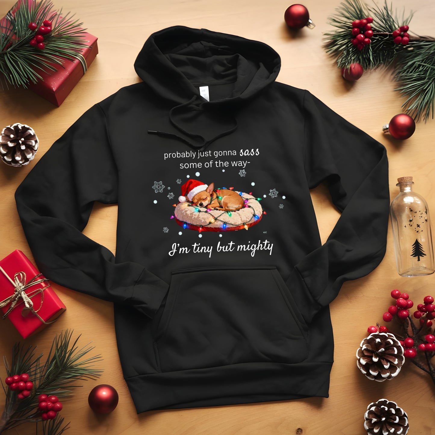 Black hoodie with Christmas-themed design on a wooden surface with holiday decorations.