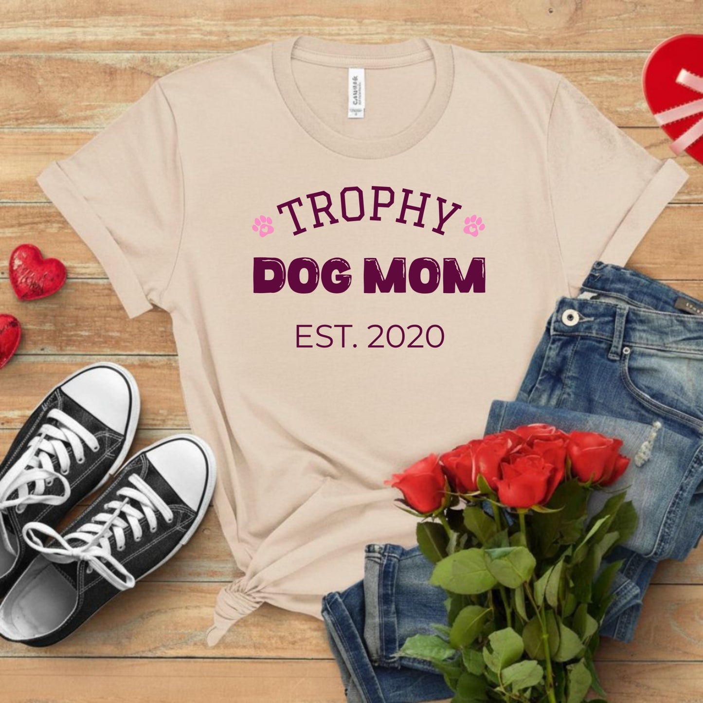 Beige t-shirt with 'Trophy Dog Mom Est. 2020' text, black and white sneakers, red roses, and denim jeans on a wooden surface.