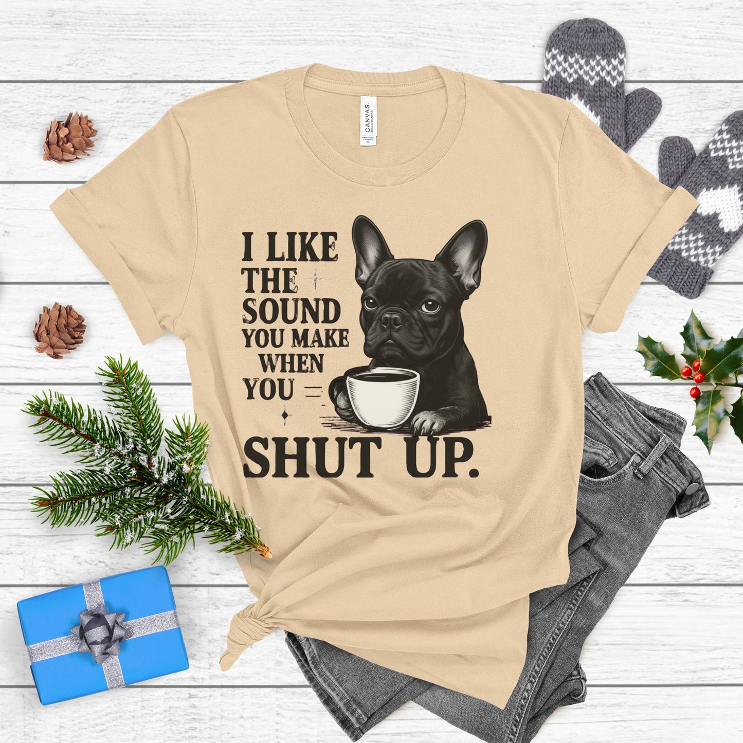 Beige t-shirt with a dog graphic and text on a wooden surface with festive decorations.