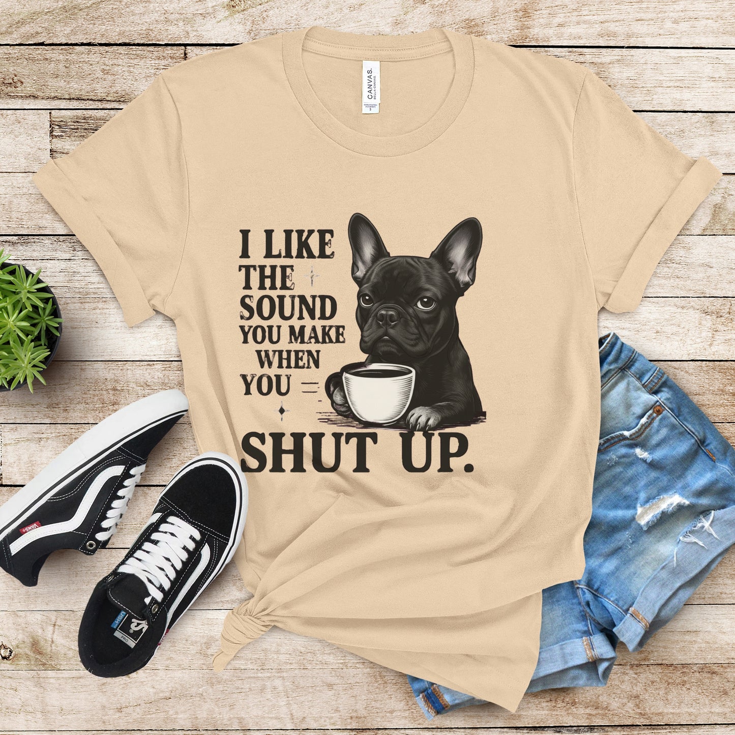 Beige t-shirt with a dog graphic and text on a wooden surface with shoes and jeans.
