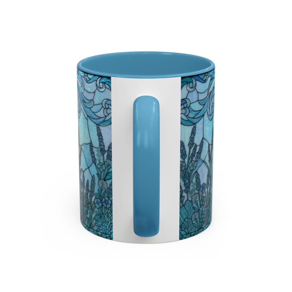 A ceramic mug with a white body, a blue handle and interior, and a blue and teal stained-glass style floral pattern on the sides, shown on a white background.