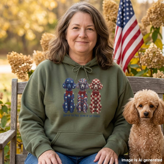 A woman sitting on a wooden bench outdoors wearing an olive green hoodie with a patriotic dog graphic, next to a small poodle and an American flag in the background.
