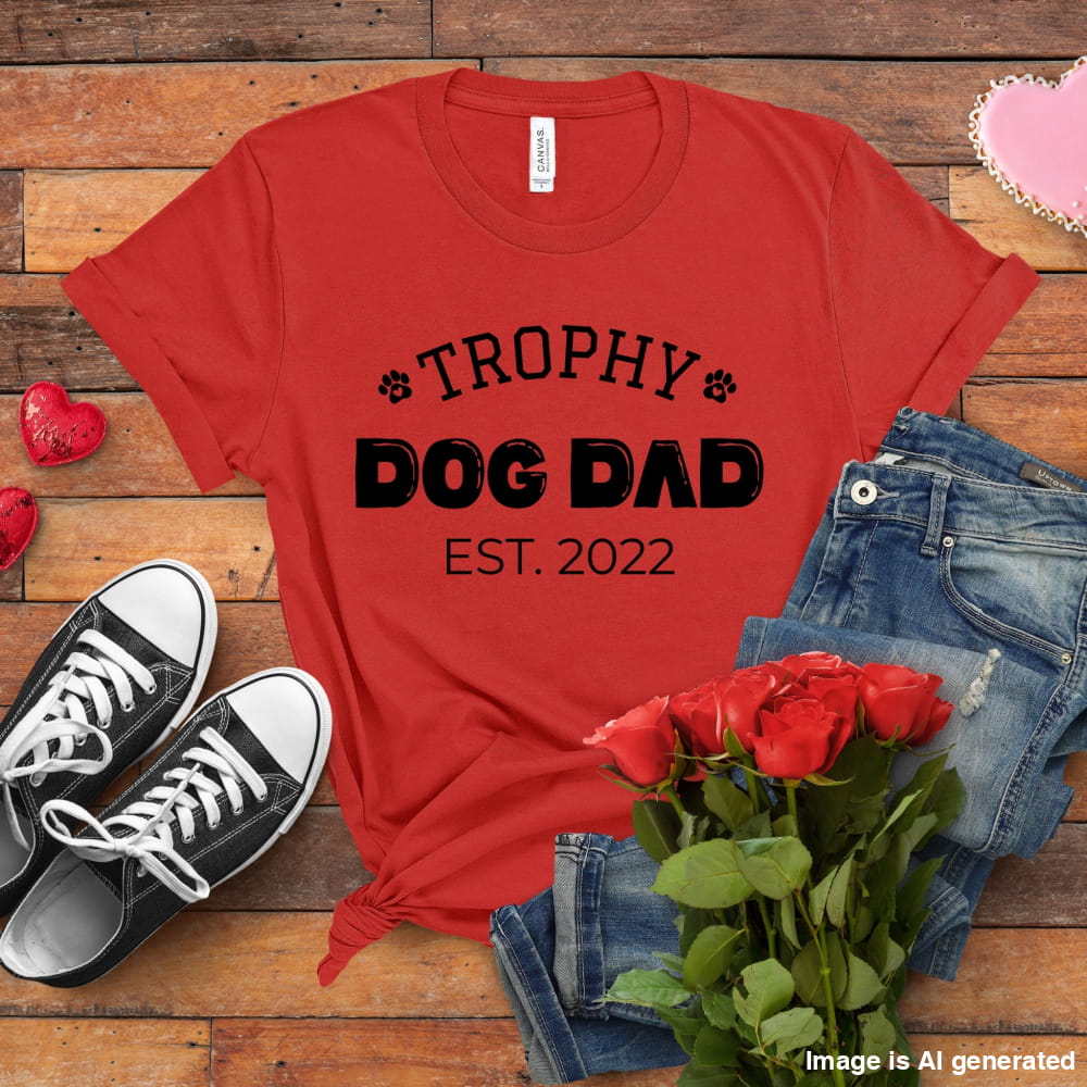 Red t-shirt with 'Trophy Dog Dad Est. 2022' text, black sneakers, red roses, and jeans on a wooden surface.