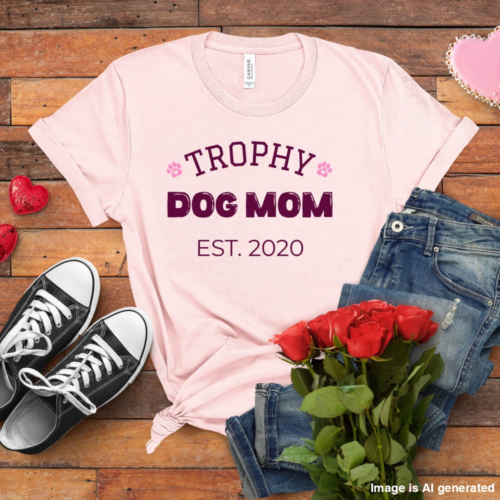 Pink t-shirt with 'Trophy Dog Mom Est. 2020' text, black sneakers, red roses, and jeans on a wooden surface.