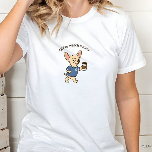 White t-shirt with a cartoon dog holding a coffee cup and text 'Off to watch soccer' on a wooden background.
