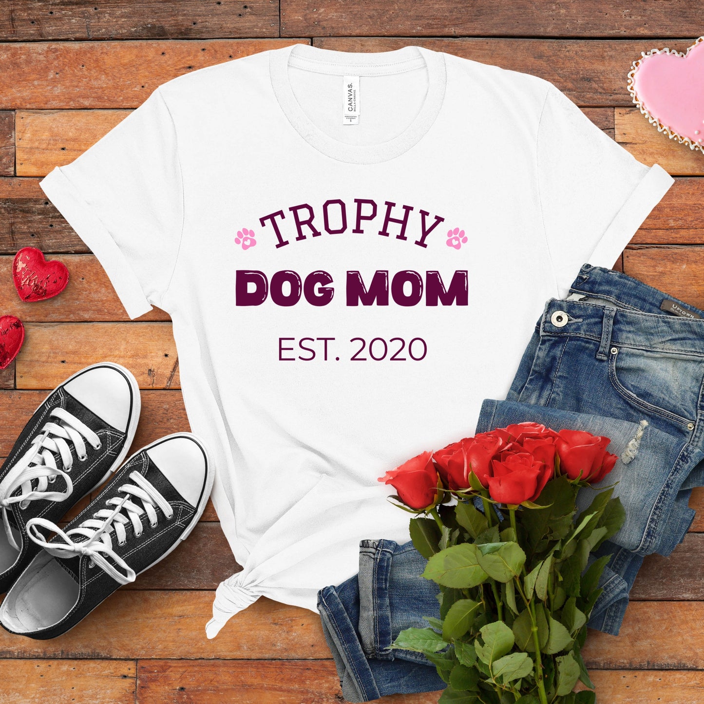 White t-shirt with 'Trophy Dog Mom Est. 2020' text, black sneakers, red roses, and jeans on a wooden surface.