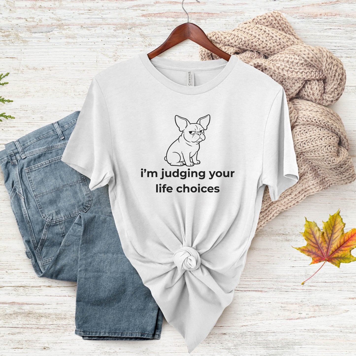 White t-shirt with a dog graphic and text, paired with jeans and a sweater on a wooden surface.