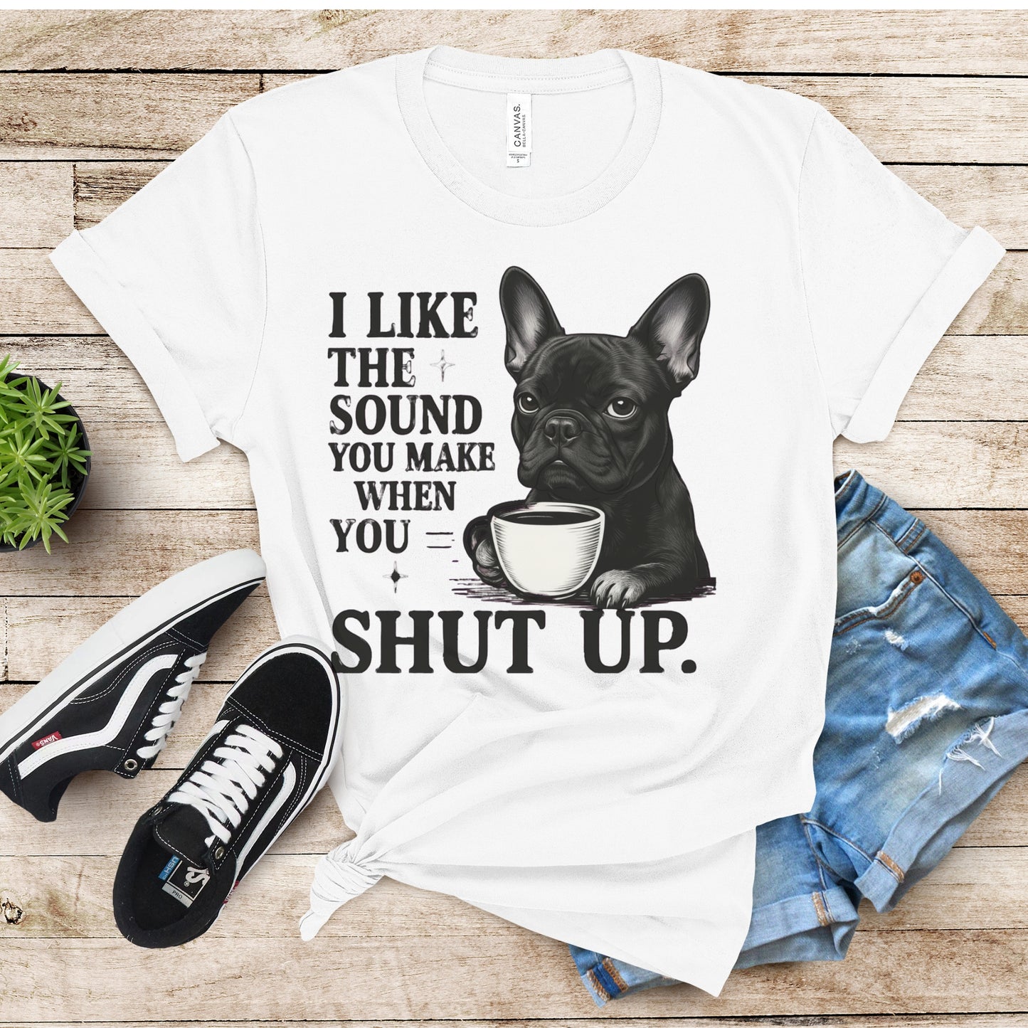 White t-shirt with a graphic of a dog holding a coffee cup and text, placed on a wooden surface with shoes and jeans.